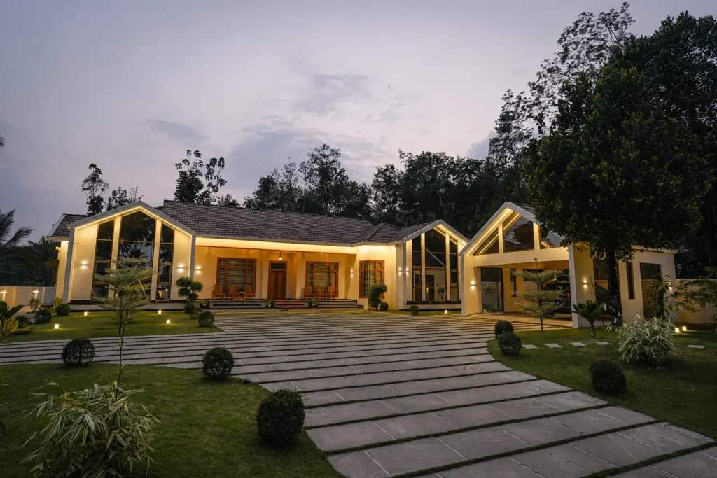 Premium modern luxury villa exterior at dusk with warm ambient lighting, sloped tiled roof, large glass facades, landscaped driveway, and contemporary residential architecture in Kerala by architects in Calicut.